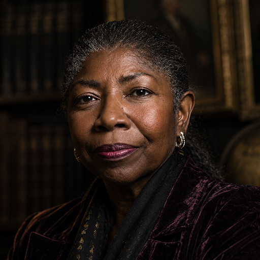 Professor Séraphine Marchetti, Chair of Marble and Requiem, photographed in lamplight before gilt-framed portraits and an old globe